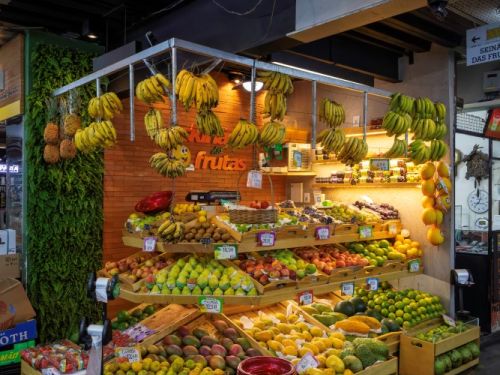 Uma banca de frutas em uma feira. A banca tem uma estrutura de madeira clara e está cheia de diferentes frutas. Em caixas e cestos no chão e sobre a bancada, há uma variedade de frutas como mangas, bananas, melões e maracujás. Algumas frutas, como cachos de bananas e abacaxis, estão penduradas em ganchos no teto.