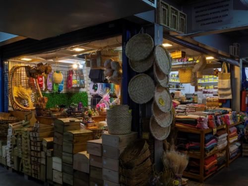 Interior de uma loja de artesanato dentro de uma feira ou mercado. A loja está repleta de produtos, a maioria feitos de palha e madeira. Há uma grande variedade de cestos de palha e tábuas redondas penduradas na frente da loja, e caixas de madeira empilhadas no chão. No interior, são visíveis bolsas coloridas, tecidos, cadeiras de palha e outros objetos de decoração.