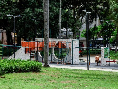 Uma vasta área verde de grama, com muitas palmeiras altas e esguias se destacando contra um céu claro. À direita, entre as árvores, um brinquedo de parque infantil com estruturas coloridas, cercado por um pequeno muro branco. Ao fundo, edifícios altos de cor clara e uma faixa de nuvens.