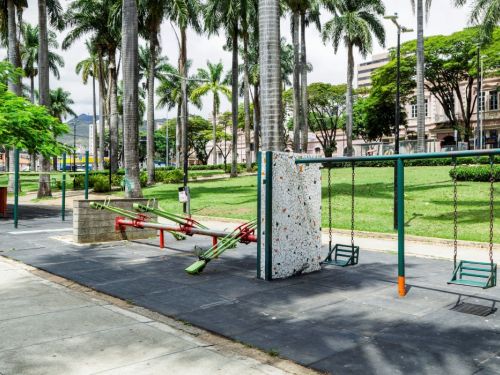Em uma área de recreação, um balanço com assentos pretos e estrutura verde-escura está à direita sobre piso emborrachado. Ao centro, um brinquedo de gangorra de metal verde e vermelho. O chão é de asfalto. Ao fundo, gramados e muitas palmeiras altas com troncos cinzentos se estendem até edifícios.