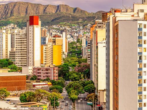 Avenida larga vista de cima, com carros e ônibus circulando entre prédios altos. Ao fundo, montanha extensa com vegetação e céu parcialmente nublado.