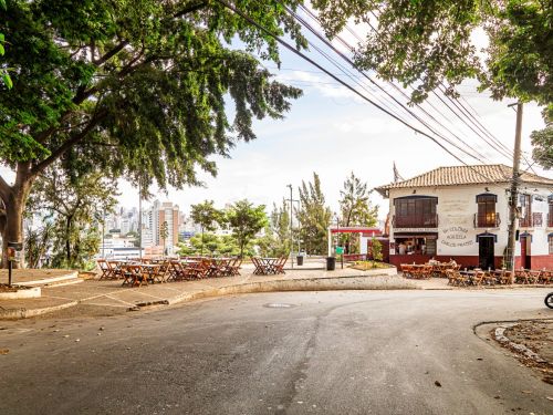 Praça pavimentada com mesas e cadeiras de madeira. À direita, prédio branco de dois andares com telhado de telhas, janelas com grades e varanda.