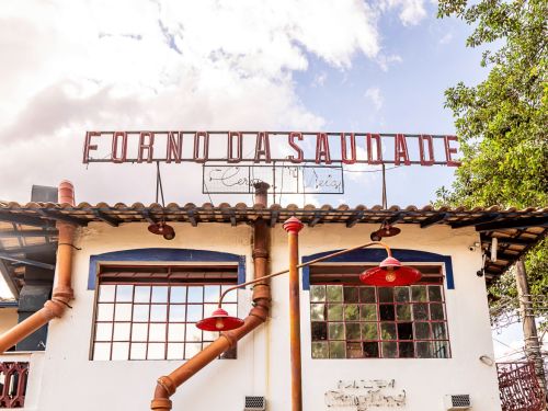 Fachada de prédio branco com telhado de telhas. Na parte superior, placa metálica vermelha com os dizeres “FORNO DA SAUDADE”. Tubulações externas ligam o teto à parede.