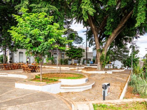 Praça com calçamento em cimento e canteiros circulares baixos. Mesas e cadeiras de madeira estão espalhadas. Árvores grandes fazem sombra no local.