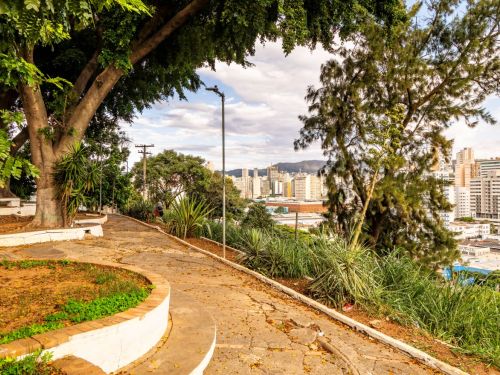 Calçada pavimentada em curvas com canteiros baixos e árvores grandes. À esquerda, vegetação rasteira e árvores. Ao fundo, prédios altos da cidade.