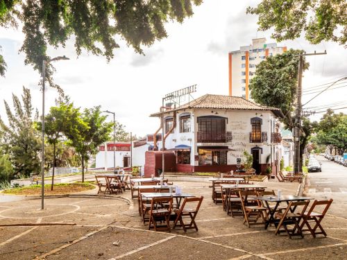Praça pavimentada com mesas e cadeiras de madeira. Ao fundo, prédio branco de dois andares, com varanda, janelas e telhado de telhas escuras.