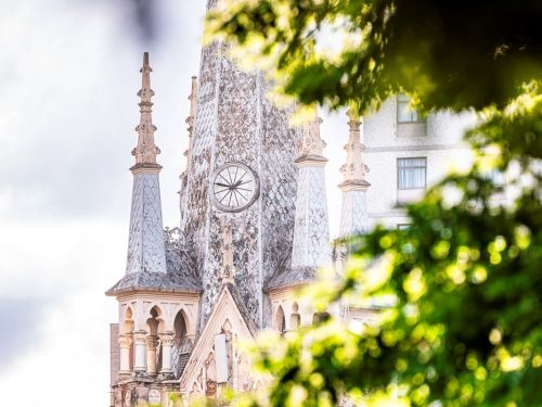 Um close na parte superior da Igreja da Boa Viagem, com a torre e um relógio. A torre é alta, de cor acinzentada, com um relógio redondo no centro. No topo, há pináculos pontiagudos e detalhes arquitetônicos. A igreja é enquadrada por folhagem verde desfocada no primeiro plano, criando uma moldura natural. 