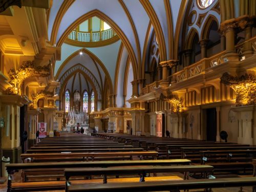 O interior da Igreja da Boa Viagem, com a nave central e as fileiras de bancos. As longas fileiras de bancos de madeira escura estão dispostas em direção ao altar, que está iluminado ao fundo. A igreja tem um teto alto com arcos brancos que se encontram no topo. As paredes e pilares são de um tom amarelo-dourado, e há uma varanda lateral sustentada por colunas. O altar, ao fundo, tem vitrais coloridos que filtram a luz.
