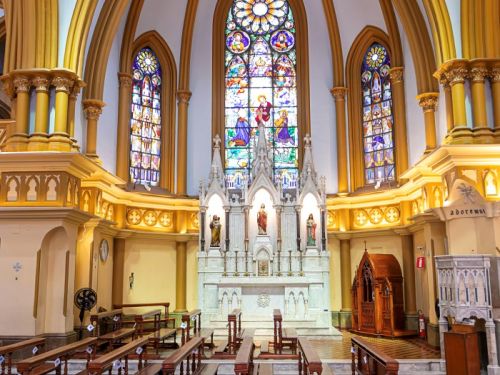 O interior da Igreja de Nossa Senhora da Boa Viagem, com os bancos de madeira e o altar. Os bancos de madeira escura estão dispostos em fileiras, de frente para um altar branco. O altar é iluminado, com estátuas de santos e uma mesa central. Acima dele, há três grandes vitrais coloridos que retratam cenas bíblicas. O teto da igreja é alto, com arcos brancos e dourados que se encontram no topo. O chão é de mármore e a luz entra pelos vitrais.