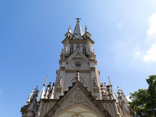 Uma vista de baixo para cima da torre central da Igreja da Boa Viagem. A torre é alta, pontiaguda e tem uma cruz no topo. A construção é de cor creme, com detalhes brancos e um telhado azul. A torre é ricamente decorada com janelas em arco, pináculos e outras esculturas. O céu é azul claro, com poucas nuvens, e a luz do sol ilumina a parte superior da igreja.