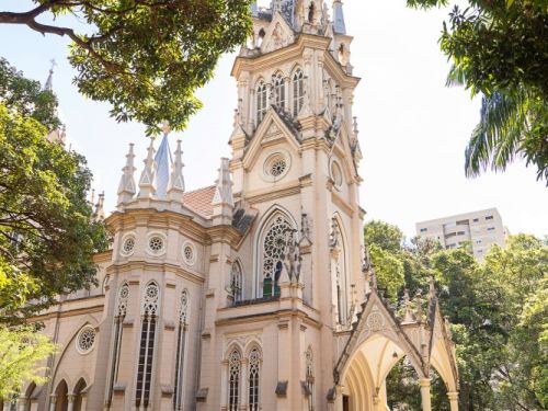 A Igreja de Nossa Senhora da Boa Viagem, uma grande construção de cor creme, vista em um dia ensolarado. A igreja tem uma arquitetura gótica, com uma alta torre central pontiaguda no topo. A fachada é rica em detalhes, com grandes janelas em arco e uma entrada coberta por um alpendre também pontiagudo. A igreja está cercada por árvores com folhagem verde escura que emolduram a cena. O chão em frente à igreja é coberto por um calçamento de paralelepípedos, e o sol brilha no alto.