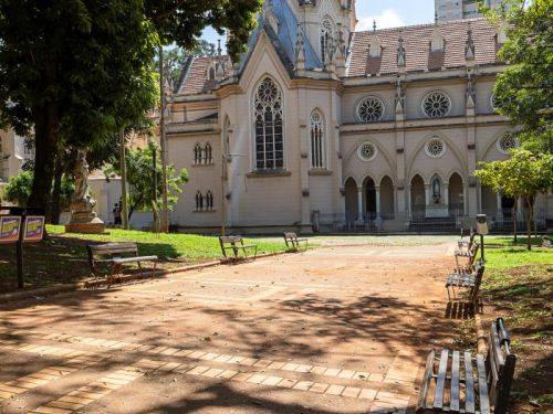 Uma vista frontal da Igreja de Nossa Senhora da Boa Viagem, uma grande construção de cor creme com arquitetura gótica. A igreja tem grandes janelas em arco e uma torre pontiaguda ao fundo. O espaço em frente à igreja é um parque ou praça, com um caminho de tijolos marrons e bancos de madeira dispostos sob a sombra das árvores. O sol da tarde bate no chão, criando um padrão de luz e sombra. 