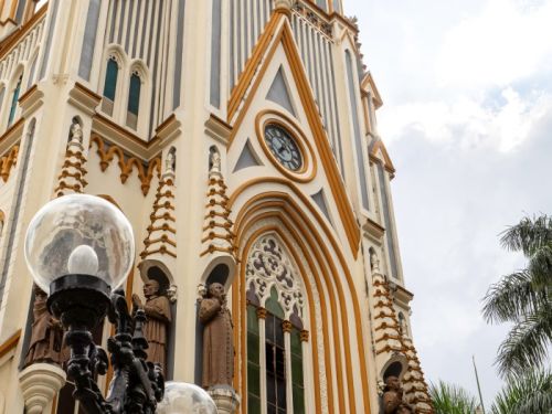 Vista de baixo para cima da fachada da Igreja de Lourdes. A construção é alta e tem uma arquitetura gótica, com uma torre central e pináculos pontiagudos. O edifício é feito de pedra clara, com listras verticais em tons de azul acinzentado e laranja-amarelado. A fachada é rica em detalhes, incluindo esculturas de santos e janelas em arco. Um grande relógio está visível no centro da torre. Em primeiro plano, no lado esquerdo, um poste de luz ornamentado com duas luminárias de vidro esféricas.