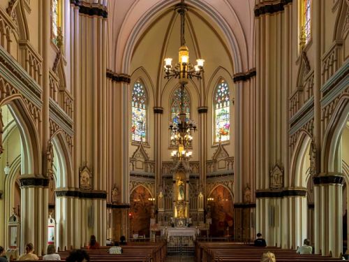 O interior da Igreja de Lourdes, uma construção de estilo gótico. O foco está na nave central, que é longa e leva até o altar ao fundo. As paredes e as colunas têm um tom creme claro e arcos altos que se encontram no teto. As fileiras de bancos de madeira escura estão dispostas de cada lado do corredor central, onde algumas pessoas estão sentadas. Grandes lustres pendurados no teto iluminam o ambiente, e vitrais coloridos nas janelas laterais filtram a luz.