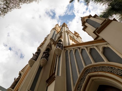 Vista de baixo para cima da fachada da Igreja de Lourdes, destacando sua arquitetura gótica. O edifício, feito de pedra clara, apresenta faixas verticais em azul acinzentado e detalhes em laranja. A fachada é adornada com esculturas de santos e pináculos pontiagudos. O céu, no fundo, é azul com nuvens brancas e densas. As folhas de árvores emolduram a cena nos cantos.