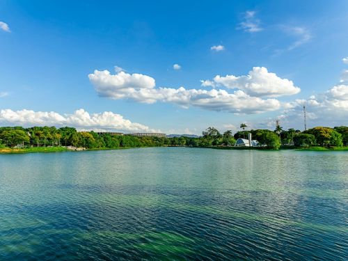 A Lagoa da Pampulha reflete um céu azul com nuvens brancas e esparsas. À esquerda, o Mineirão aparece ao fundo entre árvores verdes. À direita, a Igreja São Francisco de Assis é visível com sua estrutura arqueada branca. A água é calma, com tons de verde e azul, cercada por vegetação exuberante. O ambiente transmite tranquilidade e beleza natural.