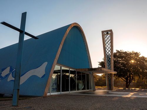 A Igreja São Francisco de Assis apresenta uma estrutura moderna com formato arqueado, revestida em azul com detalhes brancos. Um grande cruz de metal está à frente. Ao lado, uma torre estreita com grades geométricas. A entrada é composta por portas de vidro e concreto. O sol se põe ao fundo, iluminando árvores e o céu claro. O chão é de pedras irregulares.