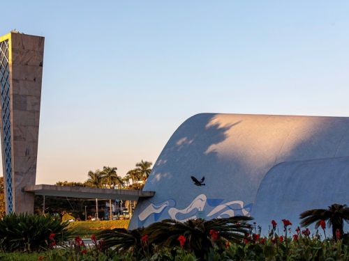 A Igreja São Francisco de Assis é destacada por sua estrutura arqueada azul e branca, com um grande campanário retangular ao lado, revestido em pedra e detalhes geométricos. À frente, plantas verdes e flores vermelhas compõem o jardim. Um pássaro voa no céu claro ao entardecer, com sombras suaves projetadas sobre a fachada.