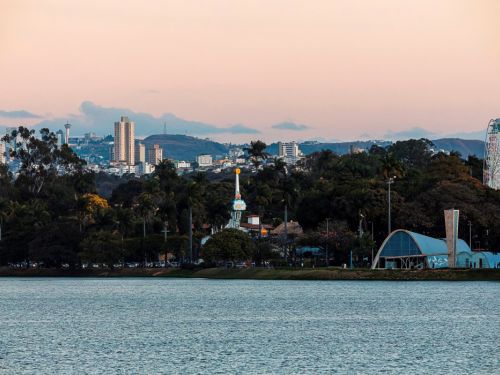 A Lagoa da Pampulha reflete o céu rosado do entardecer. À direita, a Igreja São Francisco de Assis com sua estrutura arqueada azul e campanário. Ao fundo, uma roda-gigante colorida e brinquedos de parque. Árvores densas cercam a área, com edifícios altos e montanhas ao longe. A paisagem combina natureza, arquitetura moderna e vida urbana.