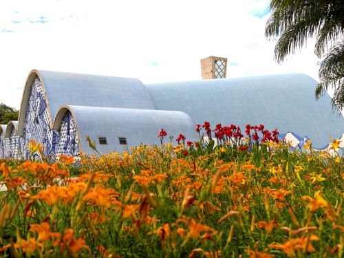 Igreja de São Francisco de Assis, em Belo Horizonte. A construção tem telhados curvos e ondulados, cobertos por azulejos azuis. Uma grande parede lateral tem um painel de azulejos azuis e brancos. Em primeiro plano, um jardim vibrante com flores laranjas e amarelas.