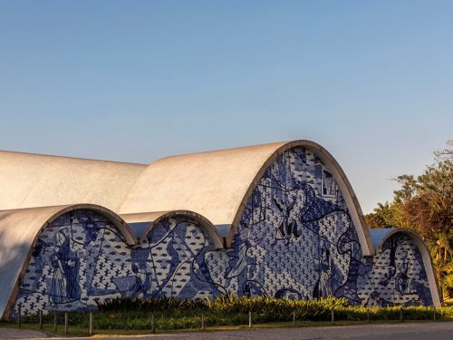 Uma vista frontal e detalhada da fachada da Igreja de São Francisco de Assis. A parede curva da igreja é coberta por um grande painel de azulejos azuis e brancos, com ilustrações de cenas religiosas. Na base da parede, há uma linha de plantas verdes. O céu está claro e azul, com árvores ao fundo.