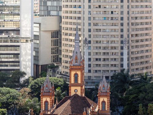 Parte superior da Igreja de São José, vista de cima e de costas. Suas três torres pontiagudas em tons de vermelho, marrom e amarelo se destacam no centro. A igreja está cercada por prédios altos, modernos e de cores claras. Árvores e vegetação verde se misturam às construções.