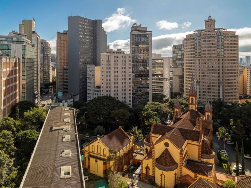 Vista aérea da Igreja de São José. Seus telhados em tons de vermelho e amarelo e suas torres pontiagudas se destacam no meio de vários edifícios altos e modernos. Árvores e vegetação verde se espalham entre as construções, sob um céu azul com nuvens brancas.