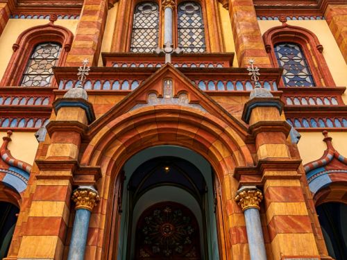 Foto de baixo para cima, foca na entrada principal da Igreja de São José. A fachada é em tons de vermelho, marrom e amarelo, com uma grande porta em arco ladeada por colunas azuis. Acima, há janelas de vitral e o relógio da torre. As portas estão abertas, e uma pessoa caminha para o interior escuro da igreja.
