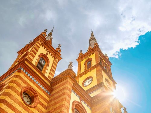 Foto tirada de baixo para cima, mostra as torres pontiagudas da Igreja de São José. As torres têm cores em vermelho-alaranjado e amarelo, com detalhes em azul. A luz do sol brilha intensamente por trás da torre central com o relógio, criando um forte clarão. O céu é azul com grandes nuvens brancas e cinzas.