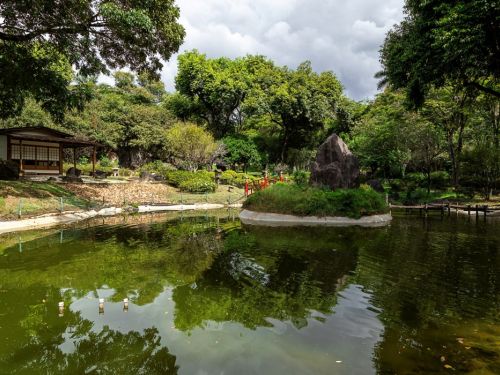 Há um lago com água calma refletindo as árvores ao redor. No centro do lago, há uma pequena ilha com uma grande pedra rodeada de vegetação. À esquerda, uma construção tradicional japonesa com portas de vidro e uma varanda coberta. O cenário é cercado por árvores verdes e um céu parcialmente nublado, criando uma atmosfera serena e natural. Pequenas pontes vermelhas conectam a ilha à margem, destacando a estética típica dos jardins japoneses.