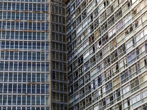 Dito pelo ChatGPT: Imagem de um edifício modernista de grande porte, visto em detalhe de sua fachada. À esquerda, a superfície plana apresenta janelas repetitivas em módulos retangulares, formando um padrão rígido e uniforme. À direita, o volume curvo do prédio exibe uma sucessão de janelas e varandas envidraçadas, muitas com cortinas ou vidros coloridos.