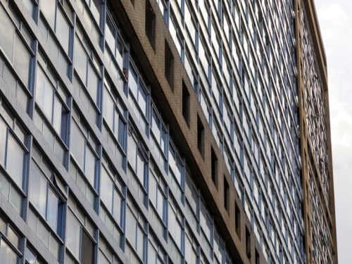 Prédio modernista, capturado em perspectiva inclinada. A fachada é composta por uma longa sequência de janelas retangulares, enquadradas por divisórias de cerâmica clara. No centro da imagem, uma faixa vertical e horizontal de revestimento marrom se destaca, o restante da superfície é envidraçada. 