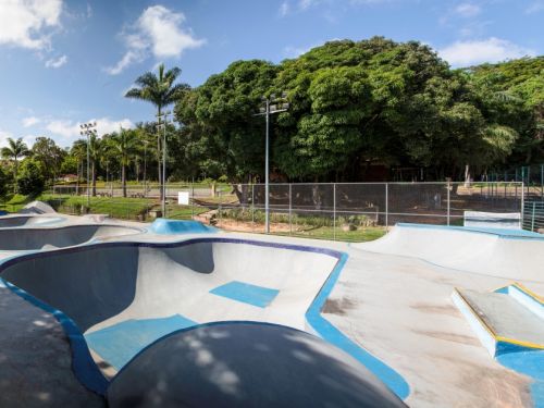 Imagem de uma pista de skate com várias rampas, bowls de concreto em diferentes tamanhos e formatos. O espaço é cercado por árvores altas e postes de iluminação. Há uma cerca metálica ao fundo.