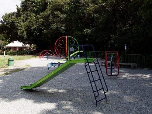 Imagem de um playground infantil ao ar livre com brinquedos coloridos de ferro. Há um escorregador verde, gangorra azul e estruturas circulares vermelhas para escalar. O chão é coberto por cascalho cinza e há árvores ao fundo.