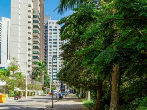 Uma longa calçada de pedras portuguesas, sombreada por árvores altas e frondosas à direita. À esquerda, corre uma rua asfaltada, e do outro lado da via, erguem-se vários edifícios residenciais altos e de arquitetura moderna, sob um céu azul com poucas nuvens brancas e altas.