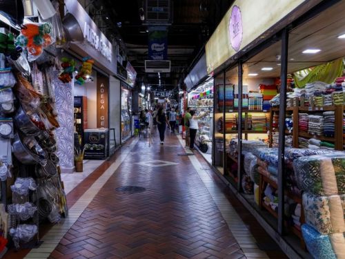 Um corredor estreito no interior do Mercado Central. O chão é de tijolos avermelhados em padrão de espinha de peixe. À esquerda, uma banca vende utensílios de cozinha. À direita, uma loja exibe rolos e pilhas de tecidos coloridos. No teto, há placas de sinalização, incluindo uma de saída. Pessoas caminham ao fundo do corredor.