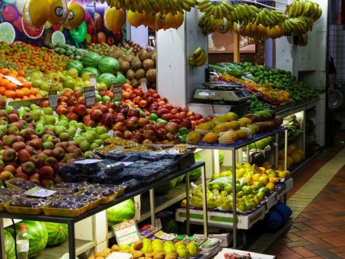 Uma banca de frutas e verduras no Mercado Central. Ela está cheia de uma grande variedade de produtos, como pilhas de maçãs vermelhas e verdes, melões, limões e bandejas de morangos. Cachos de bananas amarelas estão pendurados por cima, sob uma iluminação forte. O chão do corredor é de tijolos avermelhados.