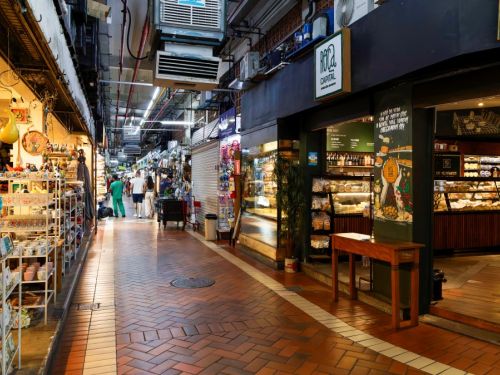 Um corredor no interior do Mercado Central. O chão é de tijolos avermelhados. À esquerda, há bancas com souvenirs e artesanato, em prateleiras brancas. À direita, uma loja com balcões de vidro que exibem pães e queijos. O teto tem tubulações e fiações visíveis. Pessoas caminham ao fundo do corredor.