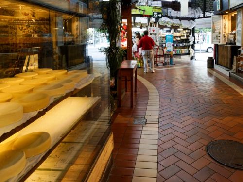 O interior de um corredor no Mercado Central. À esquerda, atrás de um grande vidro, dezenas de queijos redondos e amarelados estão empilhados em prateleiras. O chão do corredor é de tijolos avermelhados. Uma pessoa de camisa vermelha está de costas no meio do caminho, com outras lojas e pessoas visíveis ao fundo.
