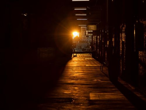Corredor escuro no Mercado Novo, BH. Uma luz dourada forte no fundo cria um caminho brilhante no chão, com reflexos e sombras alongadas. À frente, há uma grade que parece ser a origem da luz intensa. Luzes fluorescentes no teto iluminam levemente o alto do corredor.