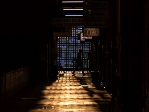 No Mercado Novo, BH, um corredor escuro é iluminado por uma luz dourada intensa vinda do fundo. Uma pessoa em silhueta caminha sobre o piso, que reflete essa luz e forma sombras alongadas. No final, uma grade quadriculada filtra a claridade. No teto, fileiras de luzes fluorescentes brancas iluminam o espaço.