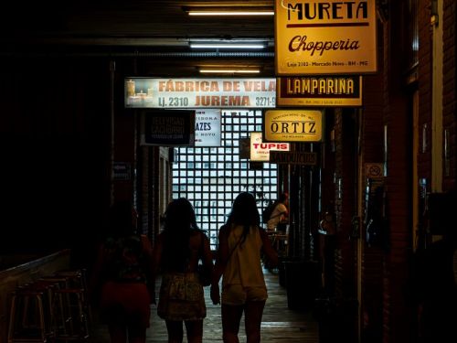 Três pessoas caminham por um corredor escuro no Mercado Novo, BH. Lojas com letreiros iluminados, como "Mureta Chopperia" e "Fábrica de Vela Jurema", pendem do teto. Luzes fluorescentes iluminam o alto e uma grade no fundo do corredor, por onde entra uma luz clara. Há banquetas à esquerda.