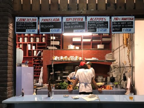 No Mercado Novo, BH, uma cozinha animada. Um balcão azul claro, com copos e garrafa, separa clientes de cozinheiros com vegetais e panelas. Acima, cinco quadros-negros pendurados listam o menu do dia e preços em "fichas", como "Carne do Dia" e "Feijoada". Prateleiras vermelhas com utensílios completam o espaço.