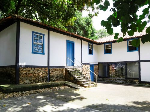Fachada lateral da casa principal do Museu Histórico Abílio Barreto. A parte inferior da construção é de pedras rústicas empilhadas, e a superior é branca, com janelas e portas azuis. Uma escadaria de pedra leva a uma porta azul. A casa está cercada por árvores, e o pátio de cimento claro tem sombras de folhagem.