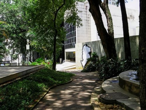 Caminho curvo de concreto em um jardim. Grandes árvores com troncos escuros estão à direita, e vegetação verde rasteira à esquerda. O chão tem sombras das árvores. O caminho leva a um prédio moderno, branco e curvo, com uma parede de vidro. Uma parede à direita tem um recorte em formato de pessoa.