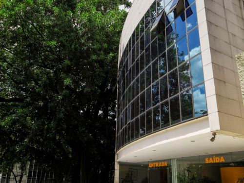 Fachada de um prédio moderno e curvo, coberto por uma grande parede de vidro que reflete o céu azul. Uma escadaria de pedra leva à entrada, onde estão os letreiros 'ENTRADA' e 'SAÍDA'. Ao lado do edifício, uma grande e frondosa árvore verde faz um contraste com a arquitetura. O céu está azul com nuvens brancas.