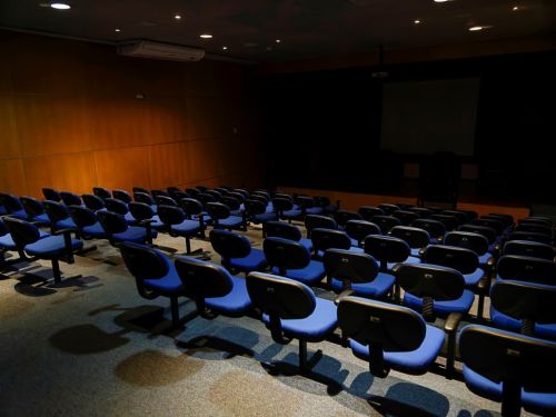 O interior de um auditório vazio. O espaço tem fileiras de cadeiras azuis escuras, com rodinhas, dispostas em duas seções voltadas para o palco. As paredes laterais são revestidas de madeira. O local é iluminado por luzes no teto, que criam sombras no chão de carpete. No palco, há uma tela escura e uma cortina preta ao fundo.