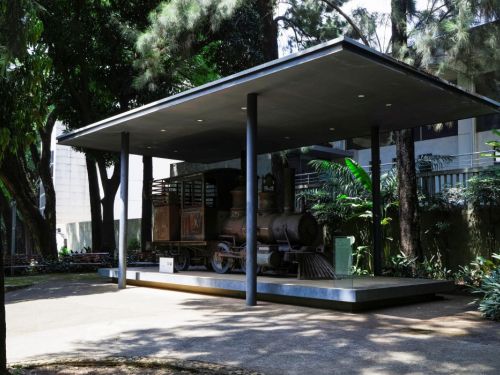 Locomotiva a vapor antiga, em exposição no Museu Histórico Abílio Barreto. A locomotiva e seu vagão, de metal escuro e enferrujado, estão sobre uma plataforma elevada, sob um telhado moderno. A peça histórica está cercada por grandes árvores e vegetação, com um prédio moderno visível ao fundo.