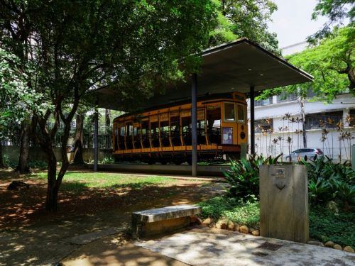 Um antigo bonde amarelo, em exposição no Museu Histórico Abílio Barreto. Ele está sob um telhado moderno, no meio de uma área com grama e muitas árvores, que criam sombras. O bonde é comprido, com grandes janelas e bancos de madeira visíveis. No primeiro plano, há um caminho de concreto e um banco de pedra.