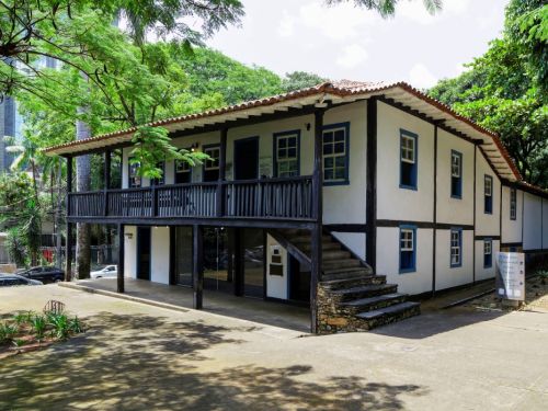 Fachada da casa principal do Museu Histórico Abílio Barreto. A casa, de dois andares, tem paredes brancas e janelas azuis. Uma varanda com cerca escura no andar de cima é acessada por uma escadaria de pedras. A construção histórica está cercada por grandes árvores e vegetação, com um prédio moderno visível ao fundo.
